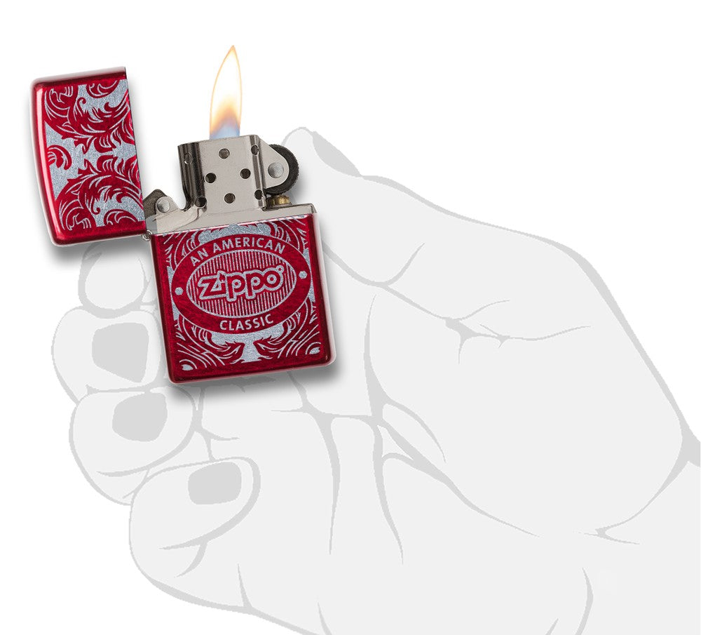 Briquet Zippo rouge  vue de face en main stylisée ouvert avec flamme entouré d’un motif en filigrane gravé au laser qui montre le logo de Zippo et de "an american classic".